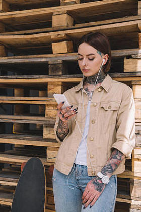 纹身时尚的女人在耳机听音乐与智能手机在滑板附近