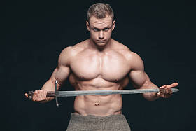 健美男子摆在黑色背景下的剑。严重赤裸的男子展示他的 mascular 身体