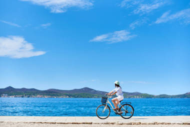 女孩騎單車在白色夏天關(guān)閉和帽子騎自行車沿石人行道上藍(lán)色波光粼粼的海水和度假村在山腳下的對(duì)岸背景。旅游和度假