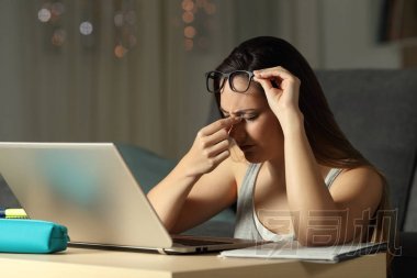 疲憊的學生在網上學習與筆記本電腦在晚上在家中遭受眼睛疲勞