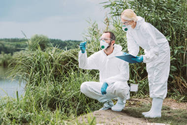 女科学家在防护服和面具写在剪贴板, 而她的男性同事检查试验瓶中的水样 