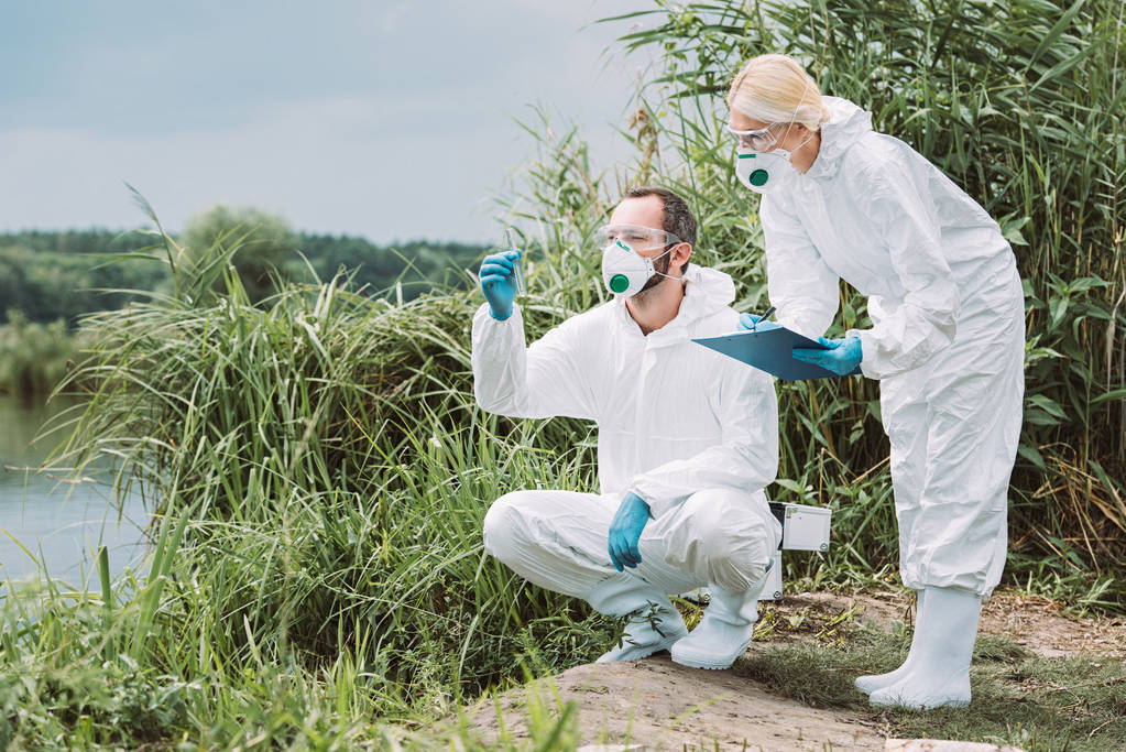女科学家在防护服和面具写在剪贴板, 而她的男性同事检查试验瓶中的水样 