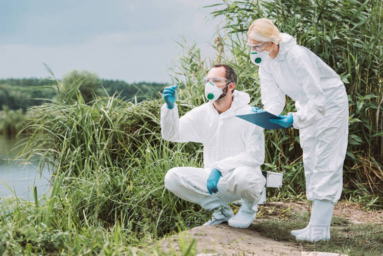 女科學(xué)家在防護(hù)服和面具寫(xiě)在剪貼板, 而她的男性同事檢查試驗(yàn)瓶中的水樣 