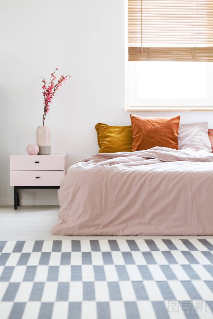 床與粉紅色的床單和橙色的墊子站在白色的臥室內(nèi)部與地毯, 窗口與木百葉窗和床邊的桌子與花花瓶