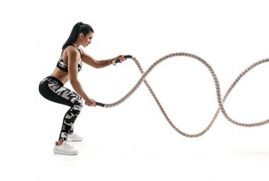 強壯的肌肉發(fā)達的女人用戰(zhàn)斗繩索工作。在白色背景下的時尚運動服中有吸引力的女人的照片。力量和動力。側面視圖