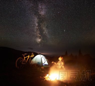 年輕女子游客在夜間露營, 在燃燒的篝火旁, 照亮的旅游帳篷, 山地自行車在驚人的美麗的夜空充滿星星和銀河。天文攝影