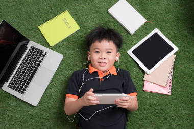 可愛的小男孩聽音樂在草地上與他的筆記本電腦, 平板電腦和筆記本電腦周圍