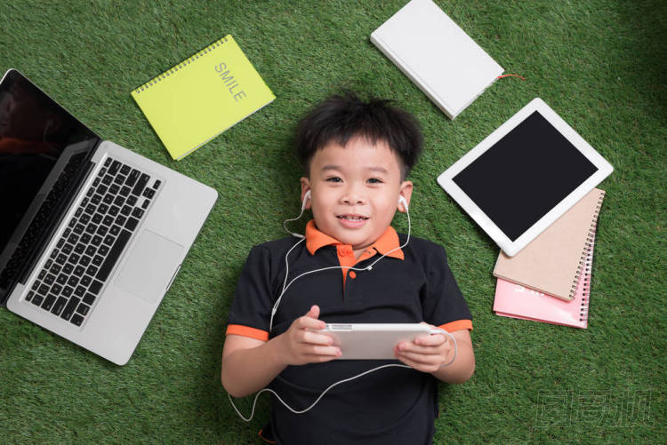 可愛的小男孩聽音樂在草地上與他的筆記本電腦, 平板電腦和筆記本電腦周圍