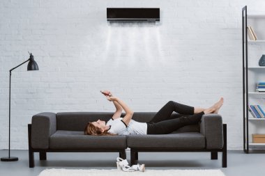 放松的年輕婦女使用智能手機在沙發(fā)下空調(diào)掛在墻上