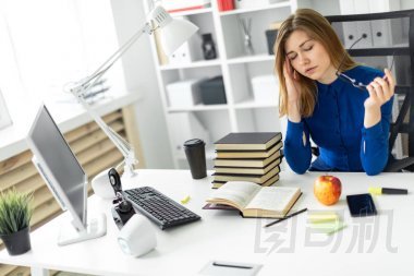 一個(gè)年輕的女孩坐在電腦桌前, 手里拿著眼鏡, 揉著眼睛。在女孩躺在一本開放的書和一個(gè)蘋果之前
