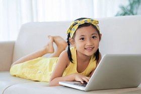 快乐的微笑的女孩躺在沙发上, 在笔记本电脑上工作