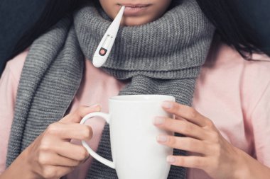 用嘴溫度計和熱飲料捧杯測量體溫的患病年輕女子的裁剪照片