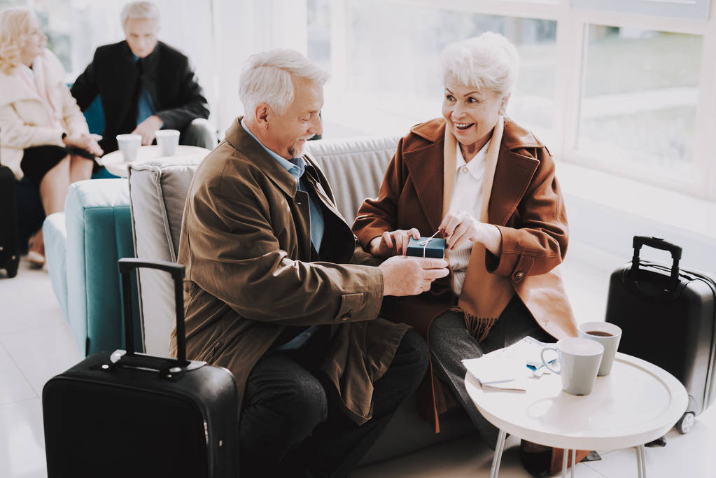 微笑的男人在机场送礼给快乐的女人。在旅行前出席。旅游理念。远航的老夫妇机场航站楼。养老金领取者的假期。携带行李的人。度假旅客 微笑的男人在机场送礼给快乐的女人。在旅行前出席。旅游理念。远航的老夫妇机场航站楼。养老金领取者的假期。携带行李的人。度假旅客