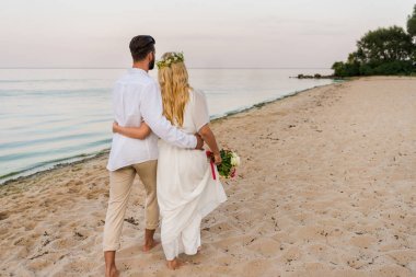 新娘和新郎的后方视图在海滩上拥抱和散步