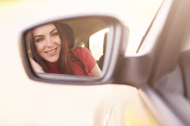 有魅力的年輕女性模特帶著溫柔的微笑, 看著汽車側(cè)面的后視鏡, 穿著紅色的衣服, 開(kāi)著汽車, 與朋友們長(zhǎng)途旅行, 享受高速。生活方式概念