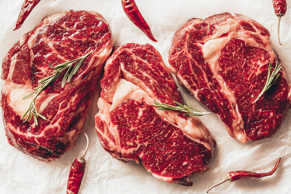 三块生肉牛排, 辣椒和迷迭香在厨房烘烤纸上的最高视图 三块生肉牛排, 辣椒和迷迭香在厨房烘烤纸上的最高视图