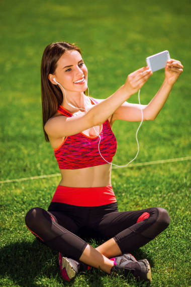 運(yùn)動禮服的年輕運(yùn)動快樂的婦女坐在草地上, 聽音樂在耳機(jī)和做自拍-照片上的手機(jī)