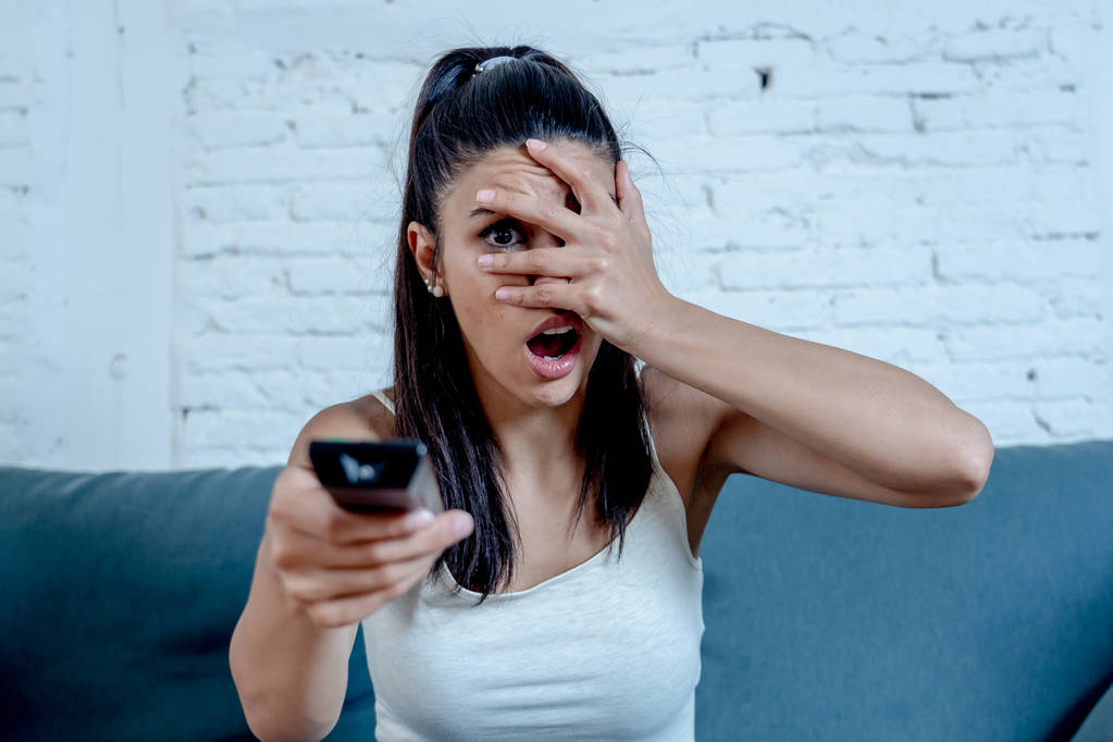 美丽的拉丁女人害怕害怕和厌恶, 而看电视恐怖电影坐在沙发上独自在客厅在家