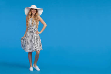 穿著白色點綴的夏日連衣裙、運動鞋和太陽帽的美麗年輕女子站著, 看著相機和微笑。在藍色背景的全長度工作室拍攝