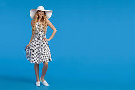 穿着白色点缀的夏日连衣裙、运动鞋和太阳帽的美丽年轻女子站着, 看着相机和微笑。在蓝色背景的全长度工作室拍摄