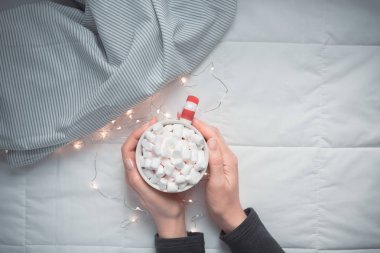 新年或圣誕節(jié)平躺頂部視圖與熱可可咖啡巧克力與棉花糖杯在婦女手圣誕節(jié)節(jié)日慶祝在床上與燈。概念博客社交媒體2019