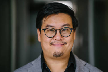 一位受過教育和中年中國亞裔 (新加坡) 男子的肖像, 穿著黑色襯衫和灰色西裝。當他凝視著相機時, 他在微笑