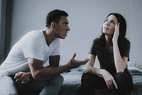 已婚夫妇坐在灰色的床上争吵。愤怒的丈夫的肖像在争吵时在妻子尖叫。女人用手牵头与人在家里有冲突。家庭关系问题