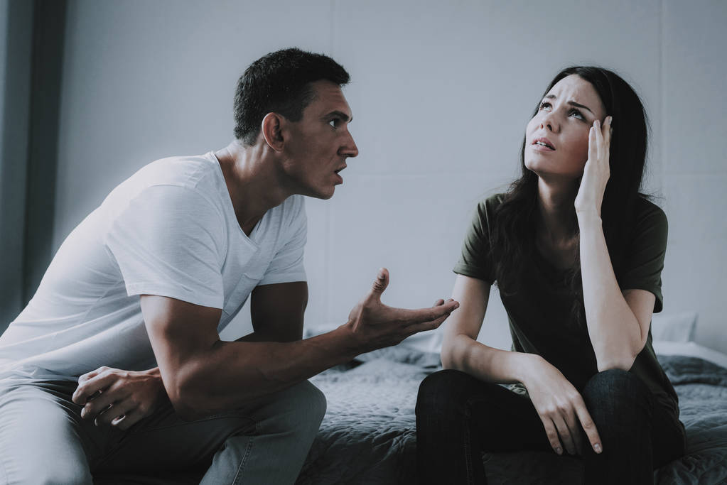 已婚夫妇坐在灰色的床上争吵。愤怒的丈夫的肖像在争吵时在妻子尖叫。女人用手牵头与人在家里有冲突。家庭关系问题