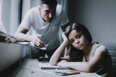 小女孩坐在桌子上, 父亲愤怒。白人黑发可爱的孩子穿着 t恤做家庭作业在记事本在家里凝视漠然在相机的肖像