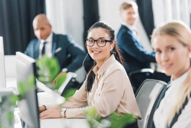 美丽的年轻女商人在眼镜和耳机微笑着对着相机, 而与台式电脑在开放空间办公室的工作