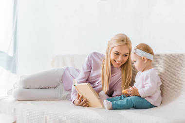年輕的微笑的母親讀書給小女兒, 而幼兒拿著餅干