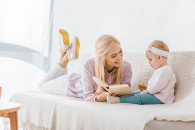 年轻快乐的母亲躺在沙发上看书给小女儿, 而幼儿拿着饼干