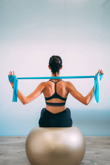 年輕的運(yùn)動迷人的婦女練習(xí)普拉提與阻力帶后面坐在健身球在白色的城市工作室。瑜伽和普拉提生活方式健康理念