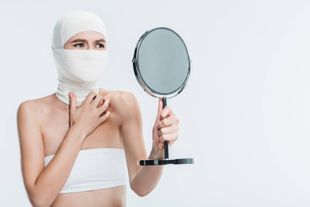 害怕的妇女整形手术后的脸上看着镜子孤立在白色 害怕的妇女整形手术后的脸上看着镜子孤立在白色