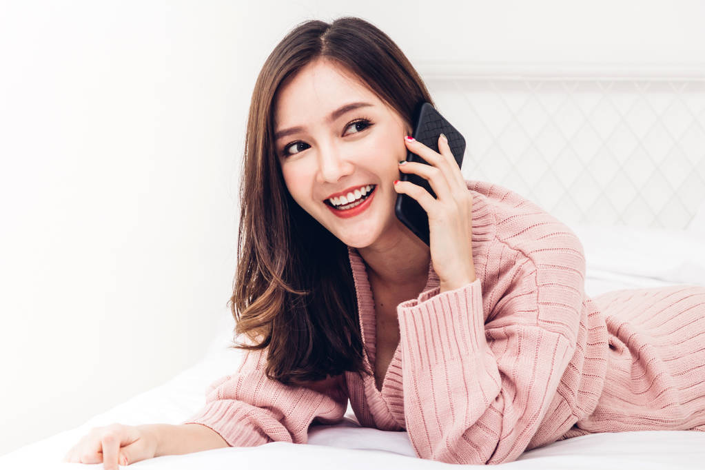 快乐的女人放松使用和交谈的智能手机在家里的床上. 技术与沟通理念