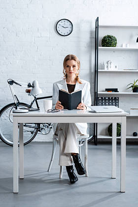 认真的女商人坐在办公桌前, 拿着笔记本, 在办公室看着相机