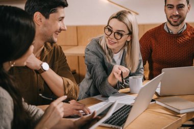 微笑的婦女在眼鏡微笑在筆記本電腦附近, 而與同事交談