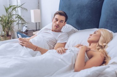 可疑的男人躺在床上與智能手機(jī)和看著睡覺(jué)的女朋友, 不信任的概念