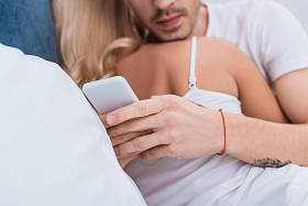 裁剪拍摄的男子使用智能手机, 而拥抱女友在床上