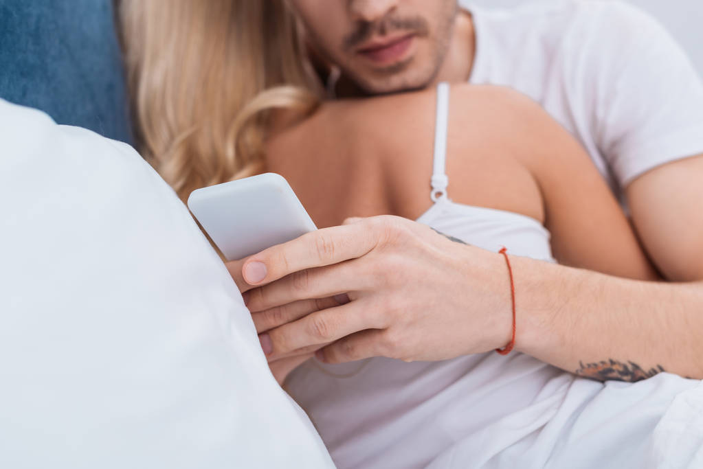 裁剪拍摄的男子使用智能手机, 而拥抱女友在床上