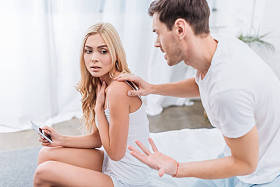 愤怒的男人触摸年轻的妻子坐在床上的智能手机, 不信任的概念