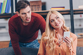 震惊的丈夫看着美丽的微笑的妻子使用智能手机在家里, 嫉妒的概念