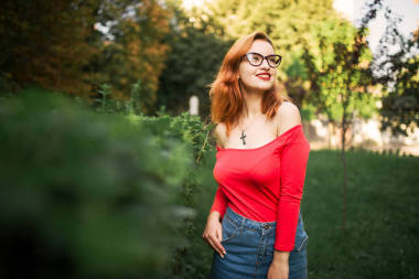 迷人的紅頭發(fā)的婦女在眼鏡, 穿紅色上衣和牛仔褲裙子擺在綠色公園