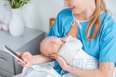 微笑的母親母乳喂養(yǎng)嬰兒和使用智能手機在病房的照片