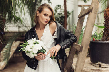 穿著黑色皮夾克和白色禮服的年輕美麗的女人手拿著小花束靠在舊的木梯上, 夢(mèng)幻般地看著旁邊舒適的溫室
