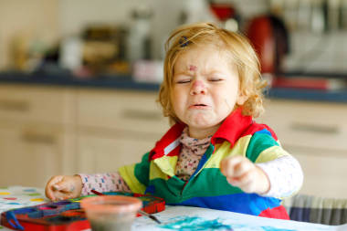 悲伤哭闹的女婴学习绘画与水的颜色。幼儿在家里画画, 用五颜六色的画笔。歇斯底里危机中的愤怒儿童