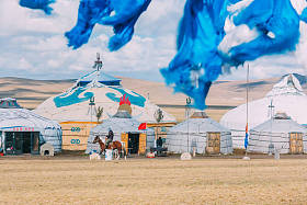 A mogolia native rides a horse in front of a group of Mongolian yurts on a grassland under a blue sky