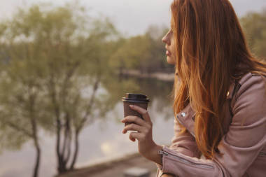 Hot red-haired woman walking in the park. It was cold and she wore a leather jacket. She drinks coffee out of a cup. Stunning sunset backdrop. Copy the space