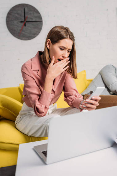 有吸引力的女人拿著智能手機和打哈欠附近的男朋友在沙發(fā)上 