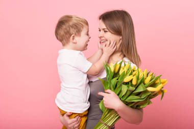 年輕的母親抱著坐在地板上的小兒子, 背景是粉紅色的。媽媽拿著一束春天的黃色花朵。關(guān)懷和人際關(guān)系以及家庭觀念。母親